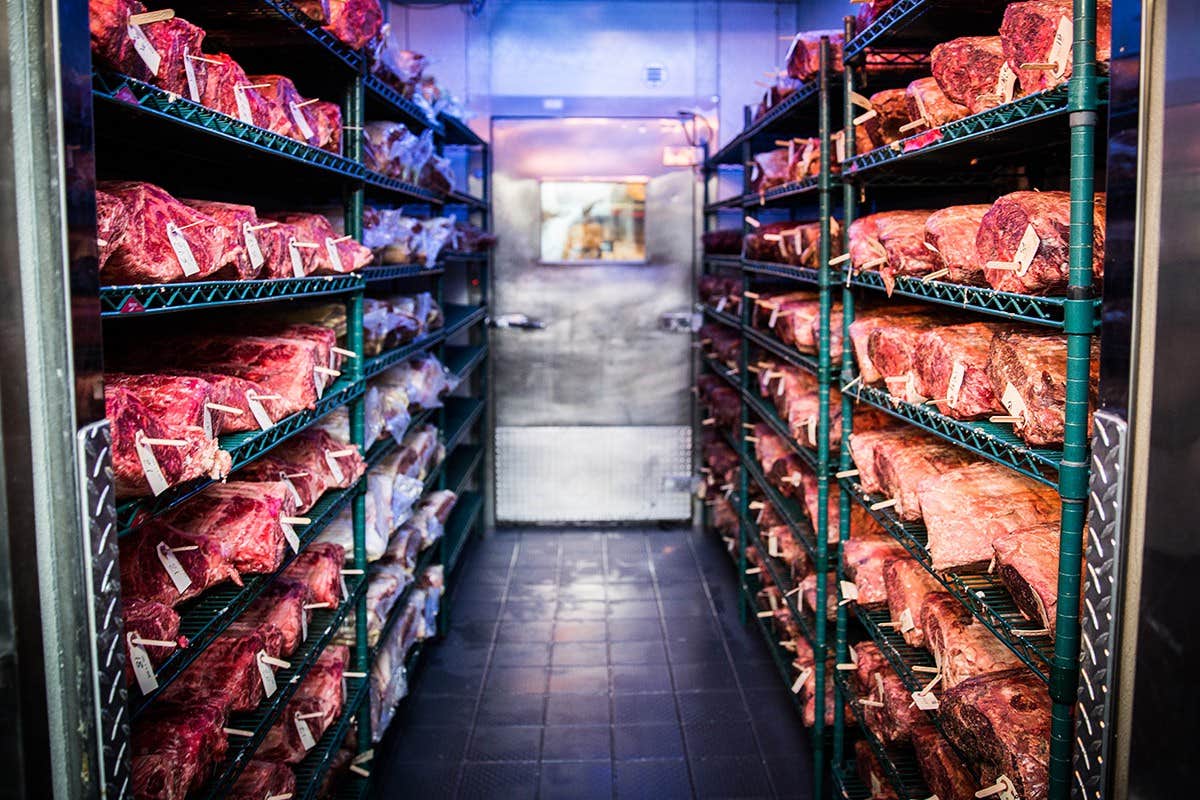 Rows of meat cuts stored on metal racks in a chilled storage room with a door at the far end.