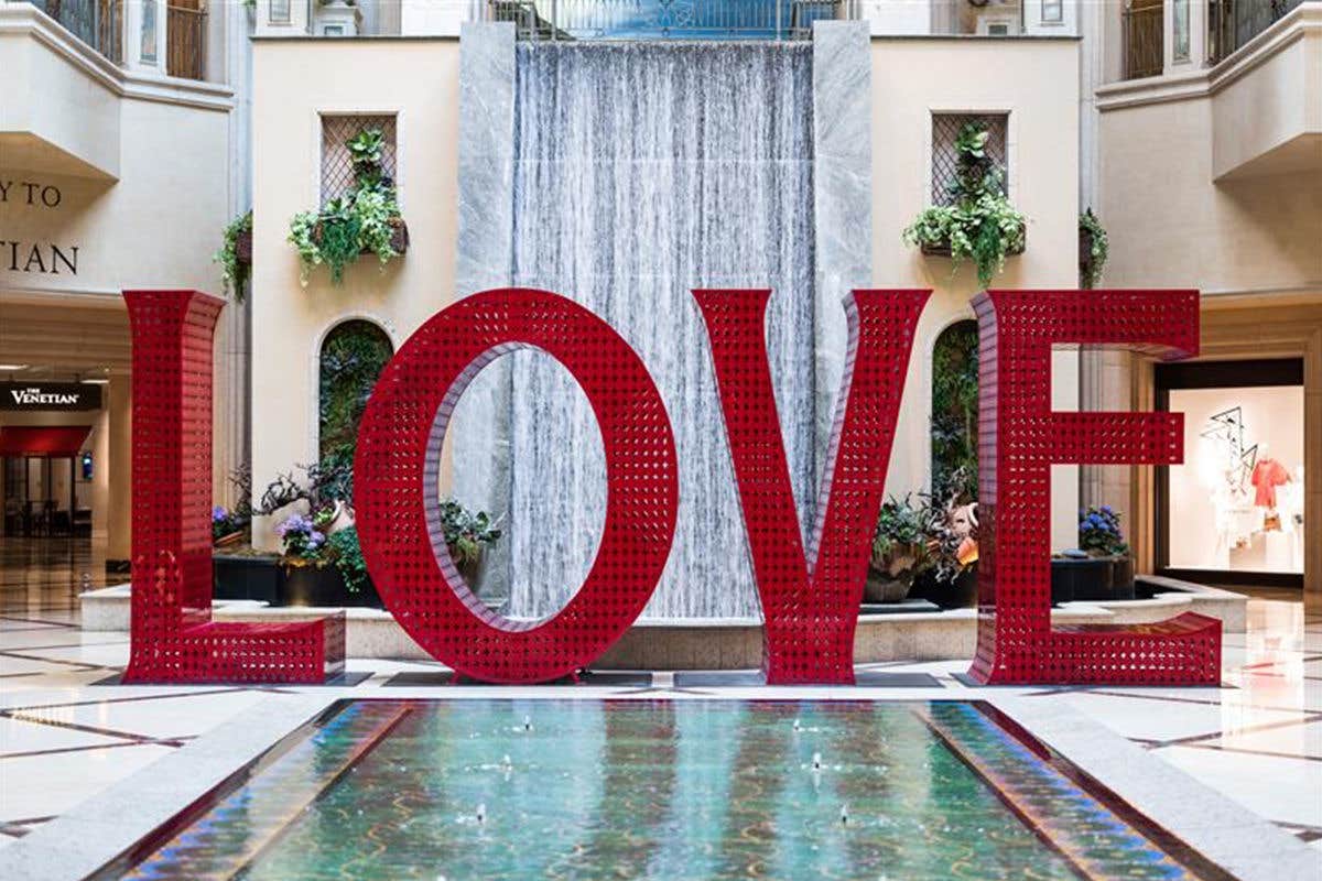 Large red letters spelling "LOVE" stand in front of an indoor waterfall, surrounded by plants and a green-tiled pool.