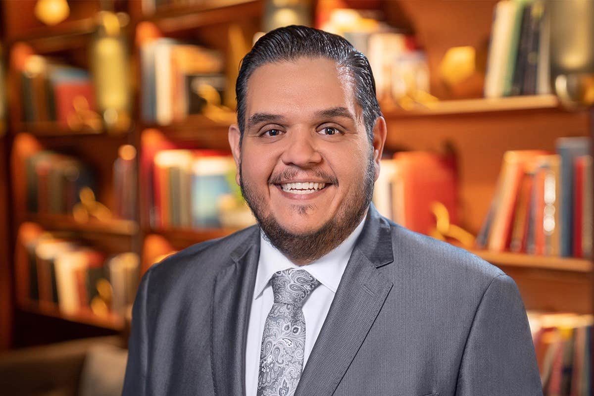 Smiling man in a gray suit and tie stands in front of a bookshelf filled with books.