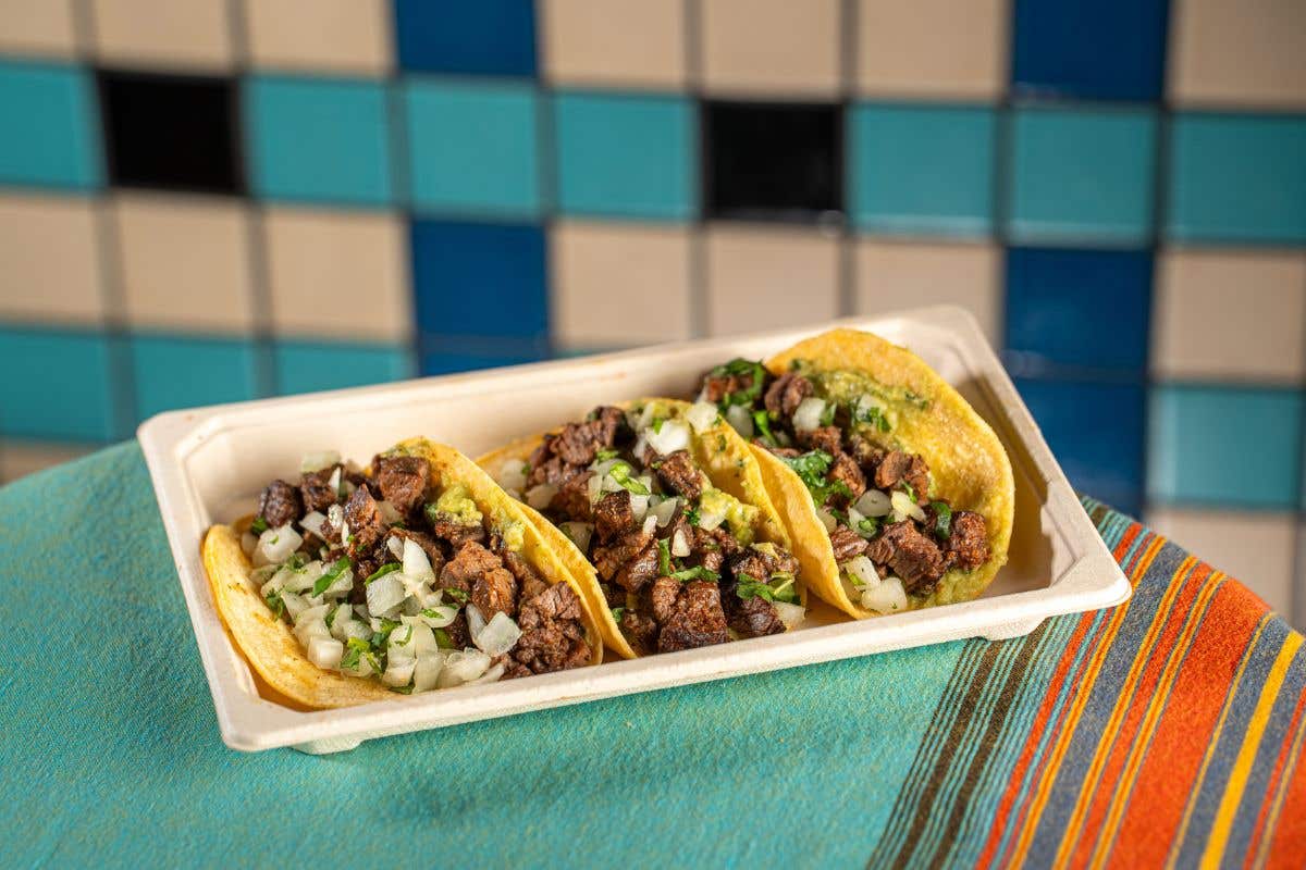 Tray of three carne asada tacos topped with onions and cilantro on a colorful tablecloth.