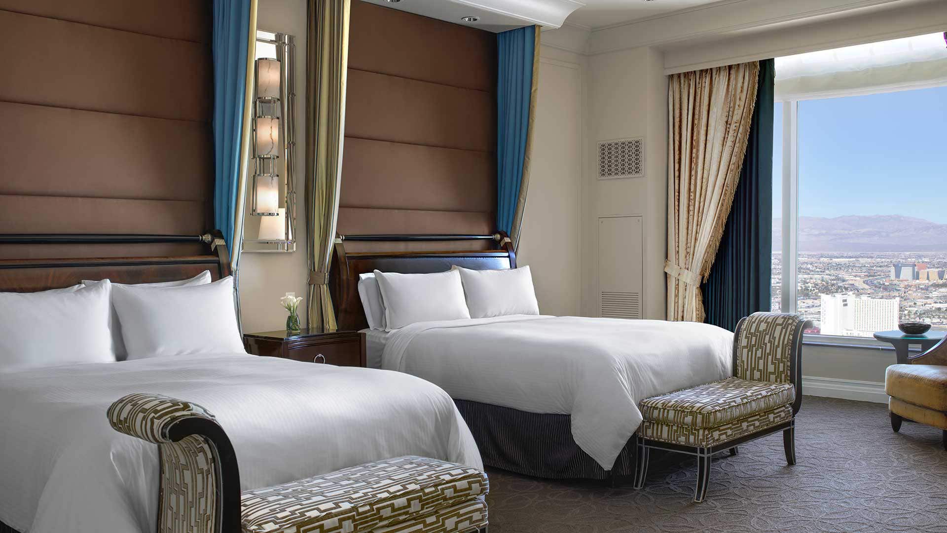 A hotel room with two Queen beds, bench at the end of each bed, and a large window to the right overlooking the city and mountains.