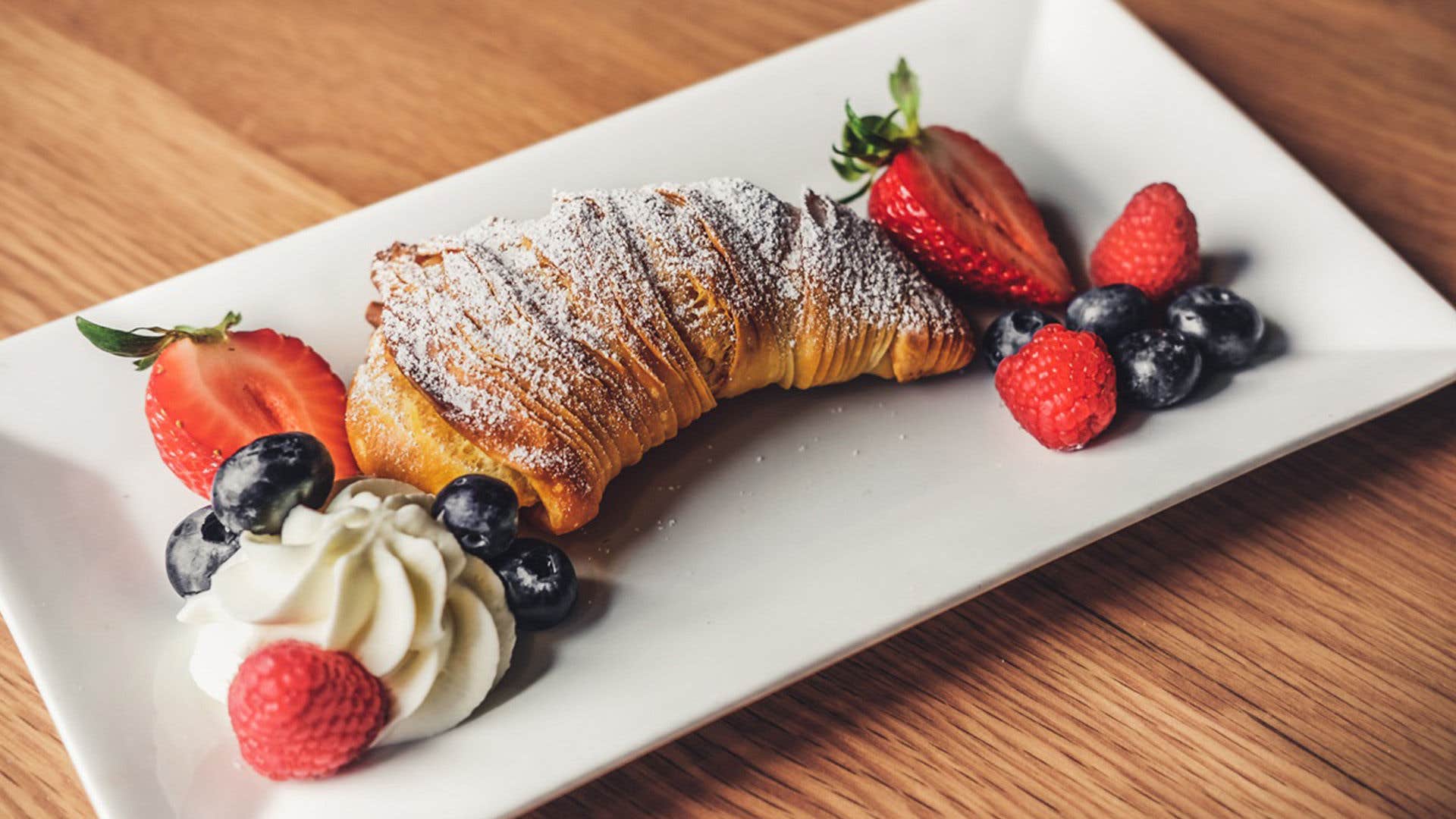 Croissant dusted with powdered sugar on a white plate, accompanied by strawberries, blueberries, raspberries, and whipped cream.