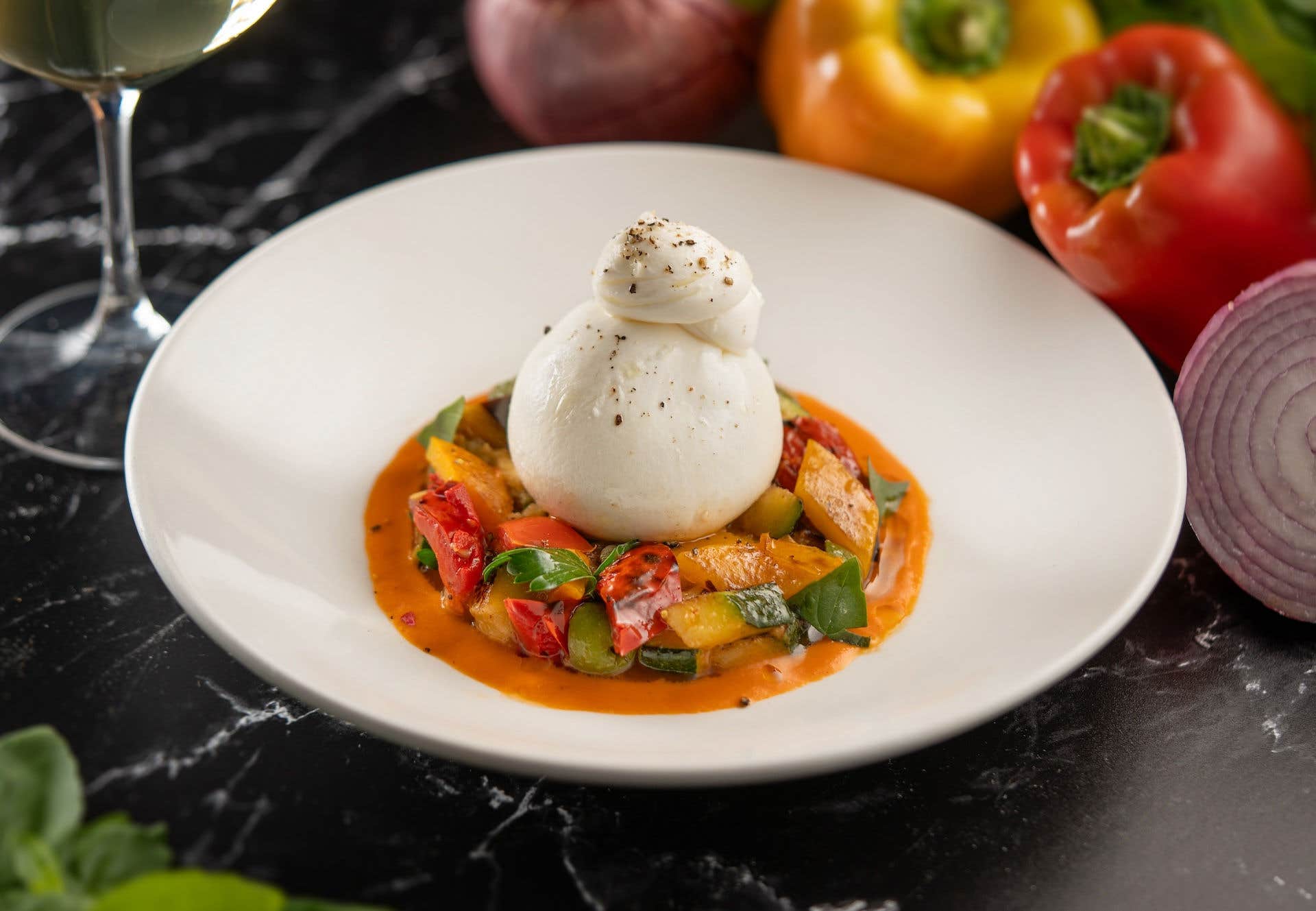 A deconstructed dish of colorful cooked vegetables topped with a generous serving of burrata cheese