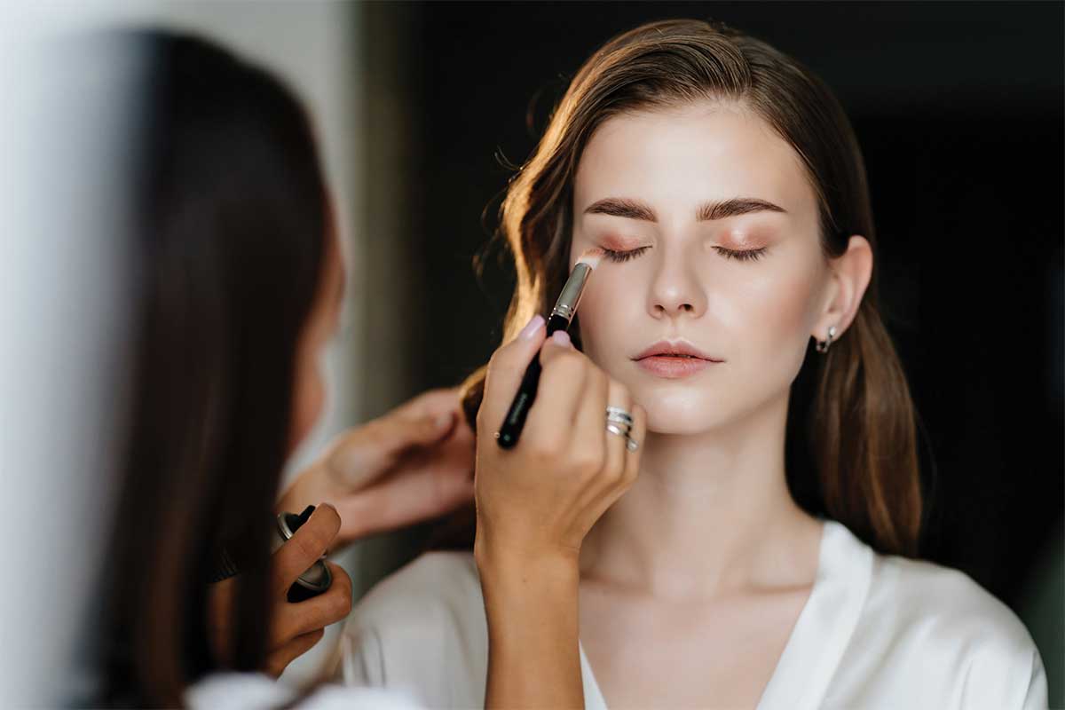 A makeup artist applies eye makeup to a woman wearing a white top, using a brush while holding cosmetic tools, in an indoor setting.