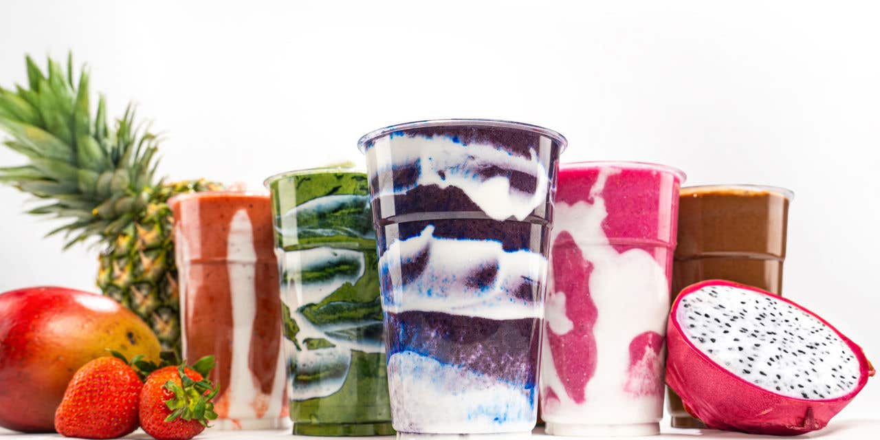 Assorted layered smoothies in clear cups with fresh pineapple, strawberries, mango, and sliced dragon fruit in the foreground.