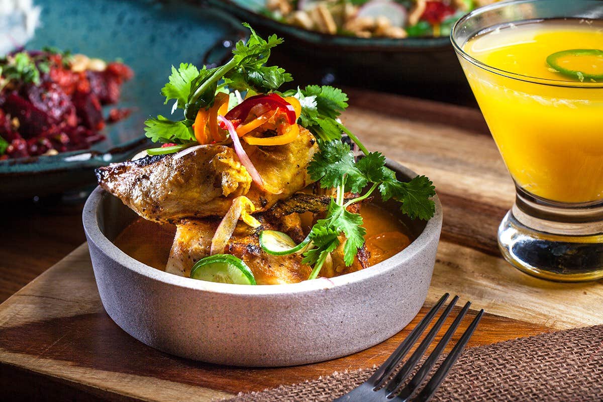 Grilled chicken topped with fresh vegetables and cilantro in a bowl beside a glass of orange beverage on a wooden table.