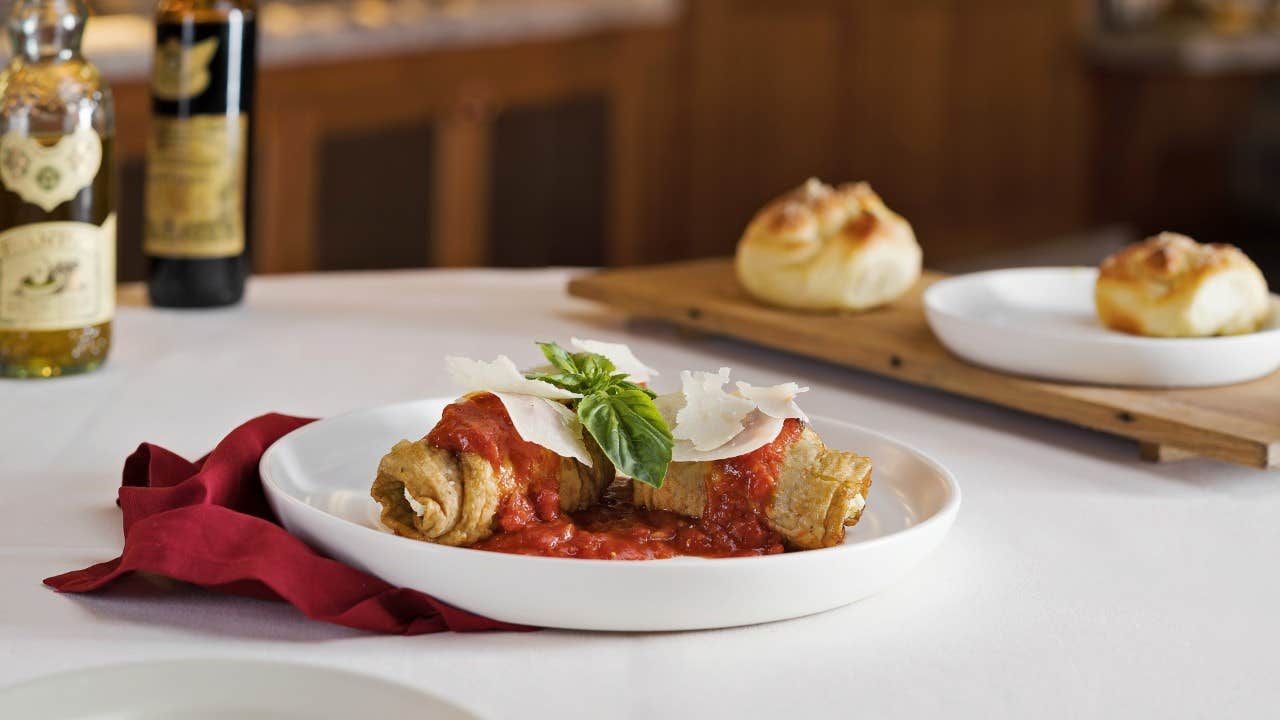 A plate of cannelloni with tomato sauce and cheese garnished with basil, accompanied by dinner rolls on a wooden board.