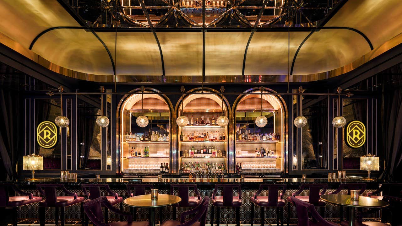 Luxurious bar with arched shelves, brass accents, and mirrored ceiling, featuring purple bar stools and round gold tables.