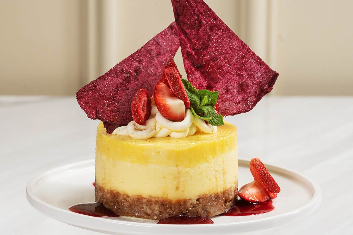 A cheesecake topped with strawberry slices, mint, cream, and red decorative pieces on a white plate.