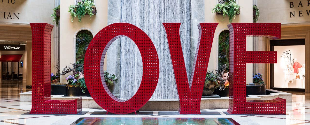 Large red "LOVE" sculpture in front of an indoor waterfall, with plants and shops in the background.