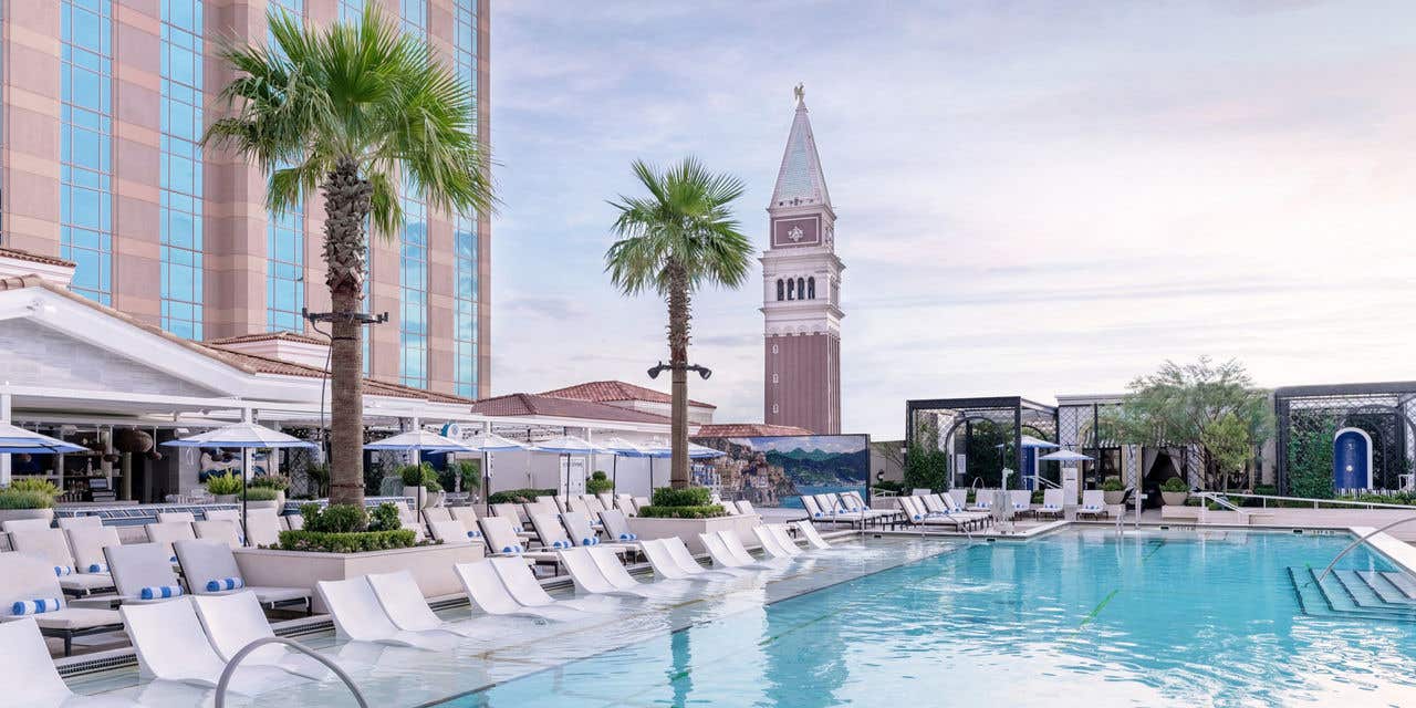 Outdoor hotel pool with two in water loungers, rolled blue and white towels, palm trees, and striped cabanas arranged symmetrically behind the pool.