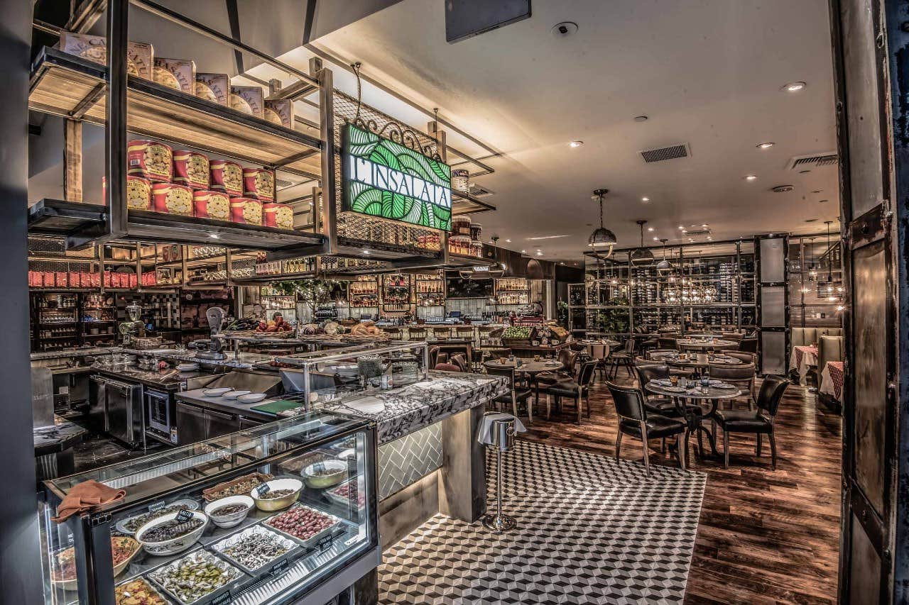 A cozy restaurant with a deli counter, shelves of food items, and tables arranged for dining under warm lighting.