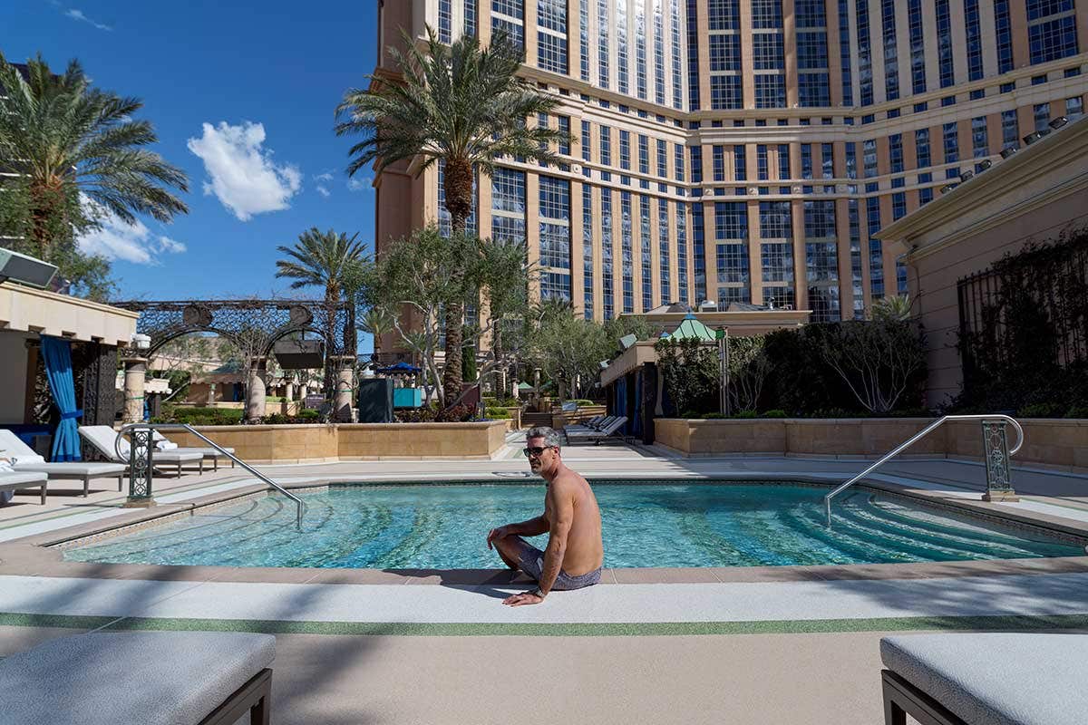 A pool with a man sitting at its edge and a hotel tower rising up in the background. 