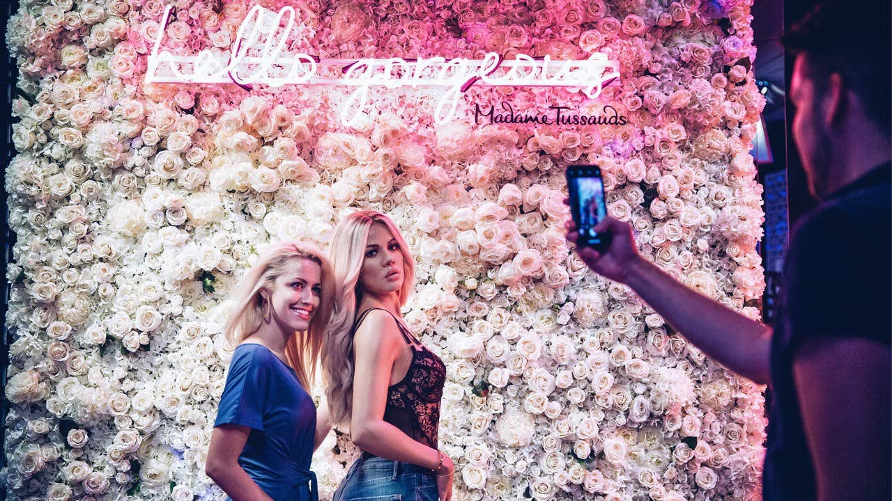 Two people pose for a photo in front of a flower wall with neon pink lights. A third person takes the picture.