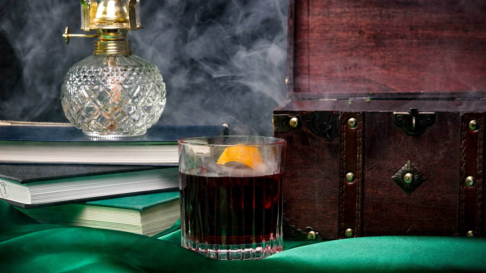 A glass of whiskey with an orange slice, books, an old lamp, and a wooden chest on a green cloth. Smoke in the background.