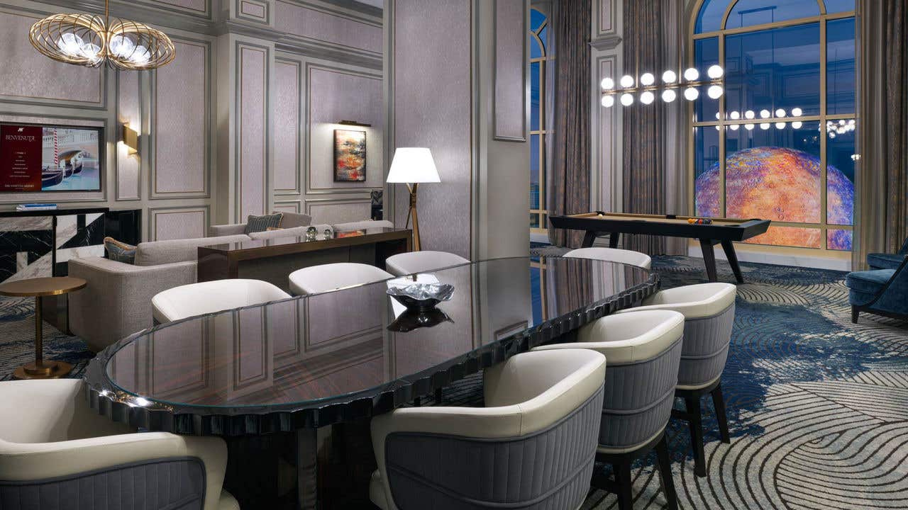 A hotel room at night with a long oval dining table with eight chairs, a couch and TV behind it to its left, a pool table behind it to its right, and tall arched windows with a view of the Sphere.