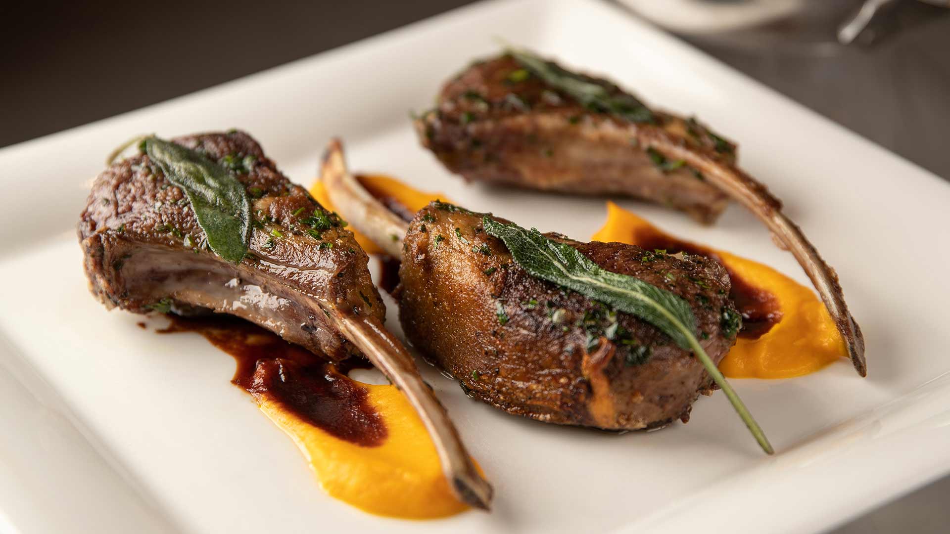 Three grilled lamb chops with herbs, served on a white plate with a butternut squash puree and a dark sauce.