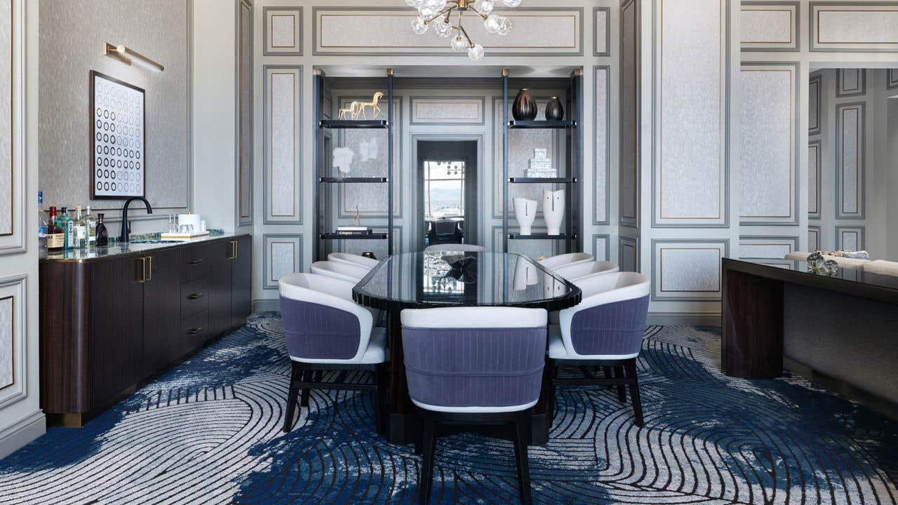 A hotel living room with a long oval dining table with eight chairs and a wet bar on the left.