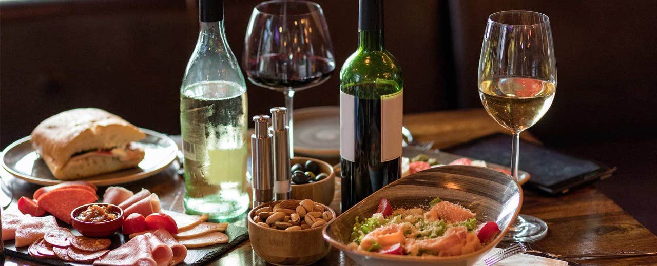 A table with two wine glasses, a bottle of white wine, a bottle of red wine, a sandwich, a salad, nuts, and sliced meats.