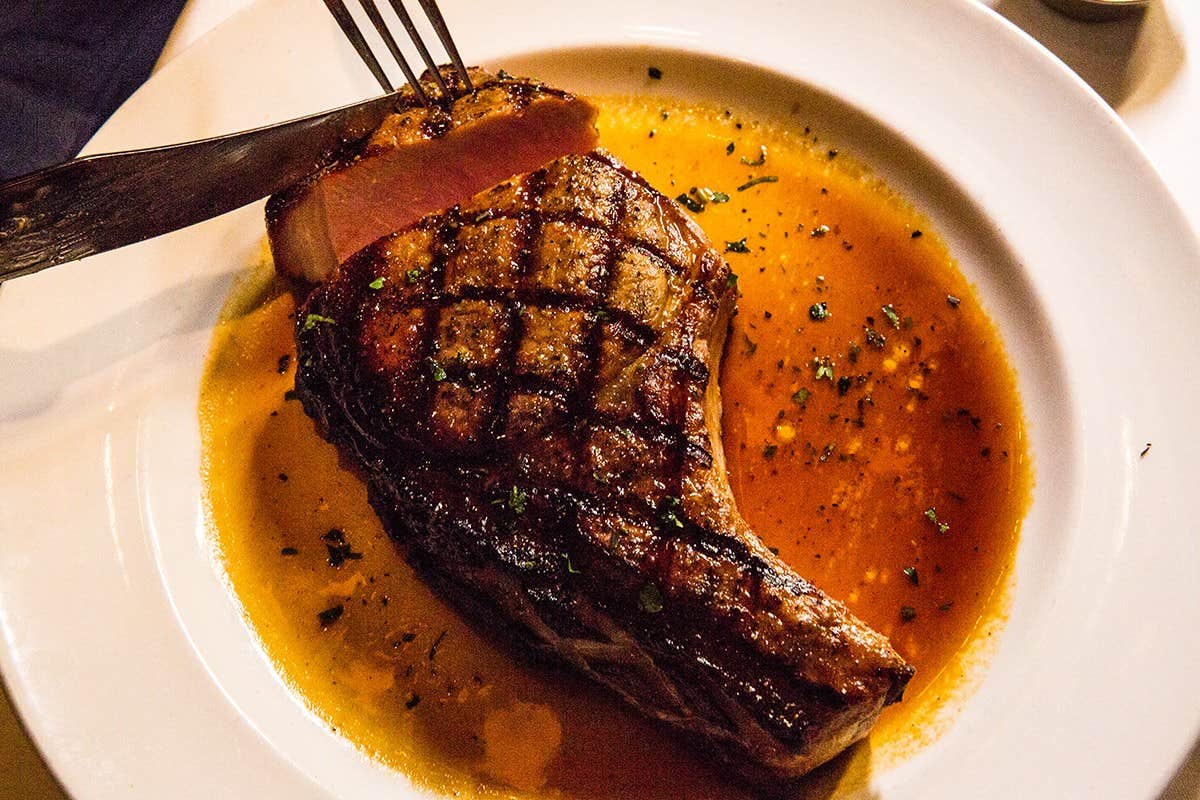 A fork cuts into a grilled steak on a white plate with sauce and garnished herbs.