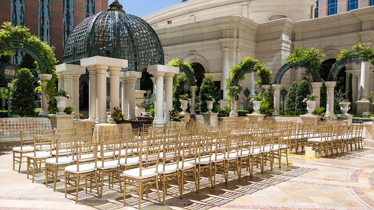 An outdoor wedding setup with gold chairs arranged in rows, surrounded by arches and greenery under a domed structure.