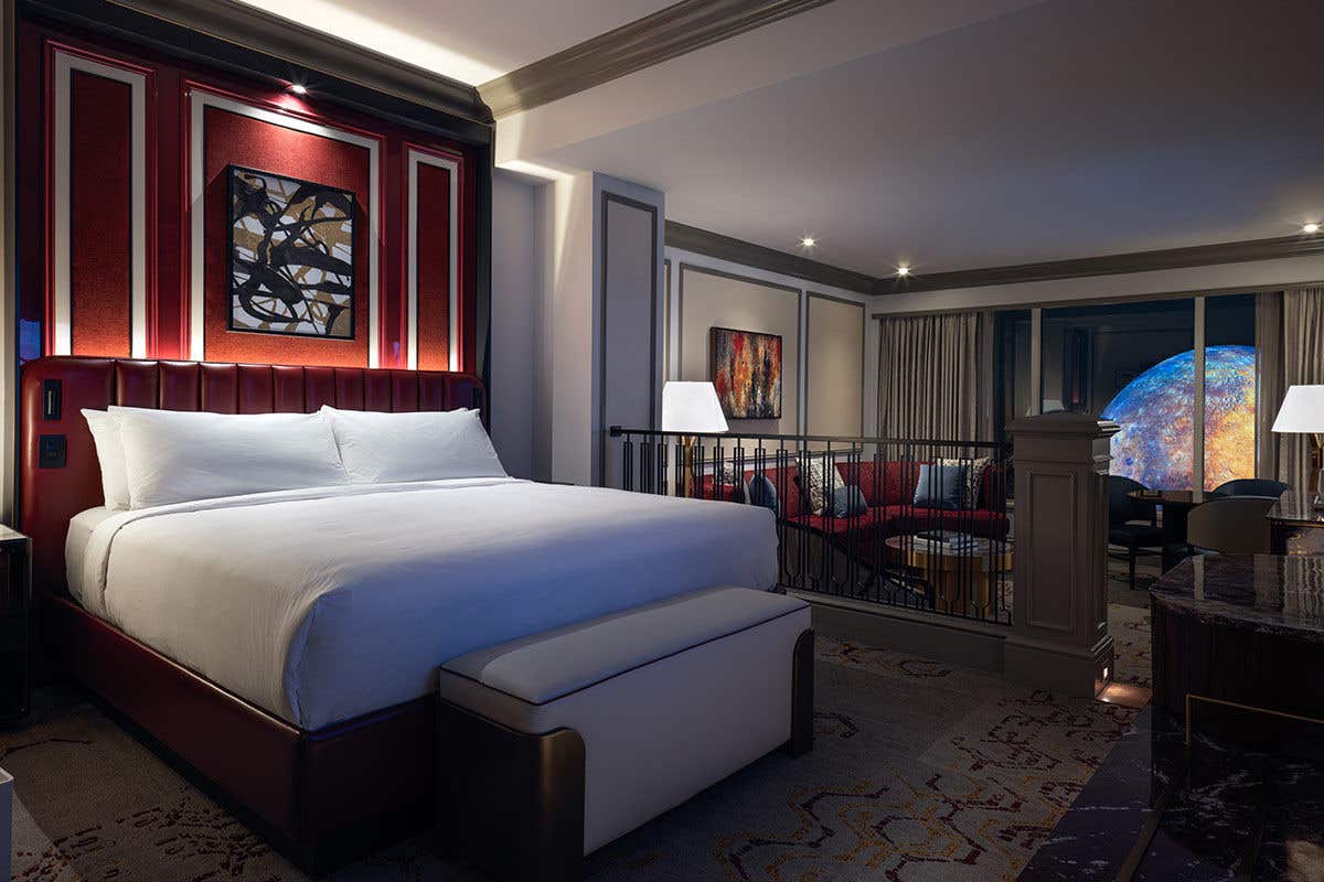 Luxury King Suite bedroom with a bench seat at the foot of the bed. In the background is a sunken living room with an armchair, couch, coffee table, dining table with chairs, lamps, and a nighttime view of Sphere at The Venetian outside the window.