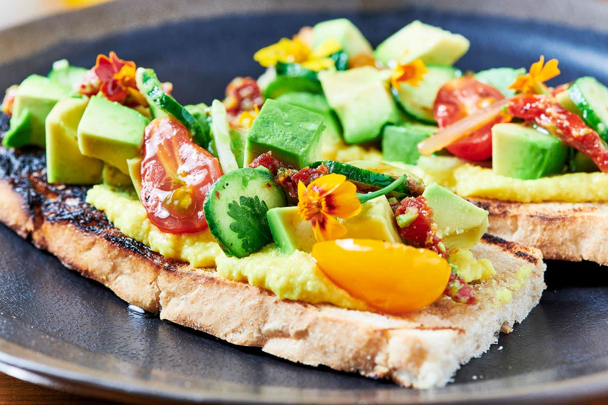 Two slices of toast topped with avocado, cherry tomatoes, cucumber, and edible flowers, served on a black plate.