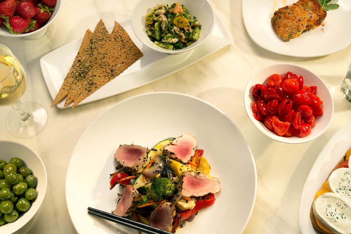 A table with various dishes: seared tuna salad, crackers, olives, tomatoes, greens, strawberries, and a glass of white wine.