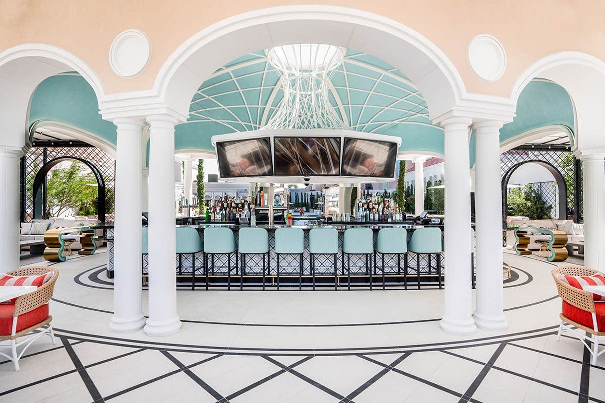 Elegant outdoor bar with light blue rooftop, white columns, and high stools, featuring a central bar counter and large TVs.