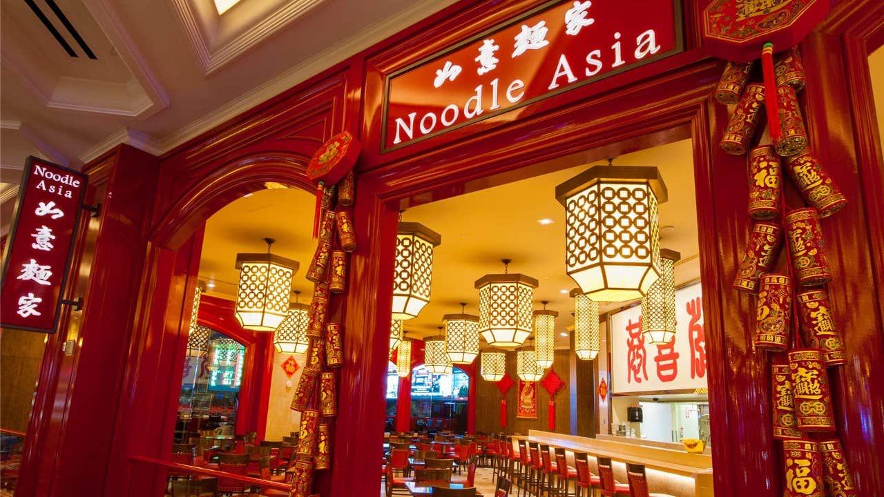 Entrance to a restaurant named "Noodle Asia," adorned with red and gold decorations, with a warmly lit interior and lanterns.