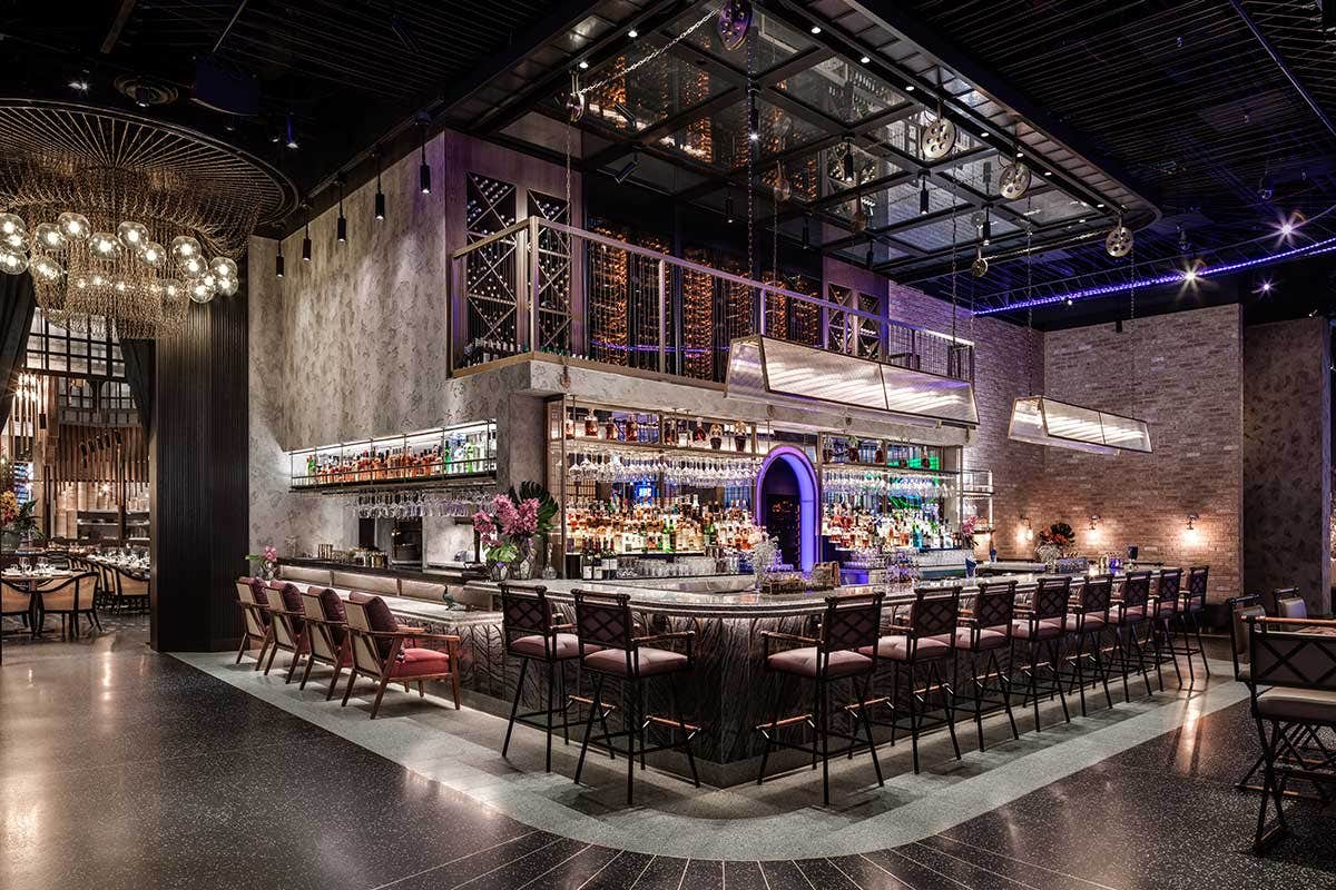 Chic bar and restaurant interior with high-top tables, an illuminated bar stocked with bottles, and modern industrial decor.