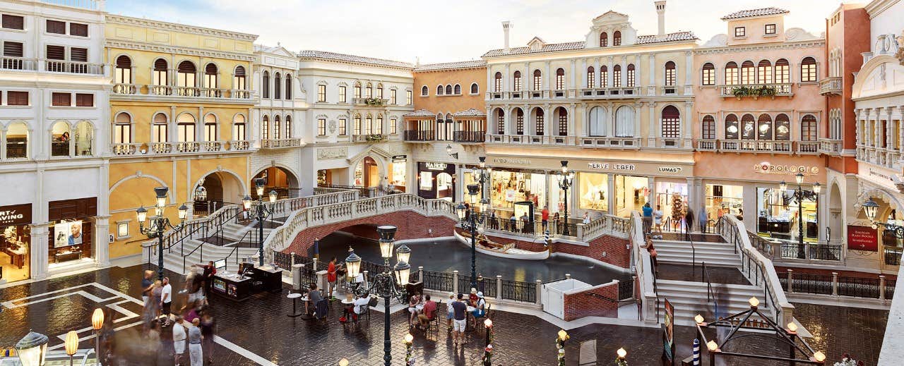 A bustling, indoor shopping mall set in a European-inspired architectural setting with a canal and pedestrian bridges.