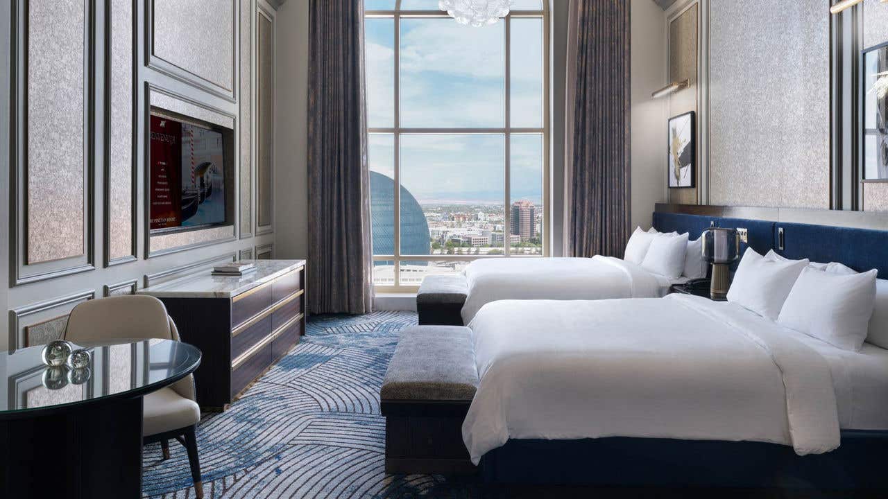 A hotel room with two queen beds, a table, a TV, a chandelier, and a tall arched window with a view of the Sphere.
