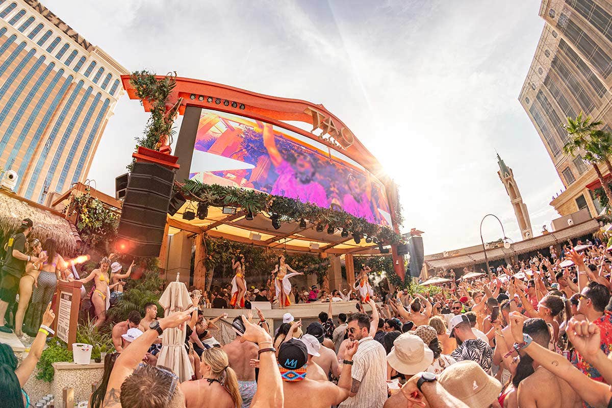 Crowd enjoying a lively outdoor pool party with a large stage and screen under bright sunlight.