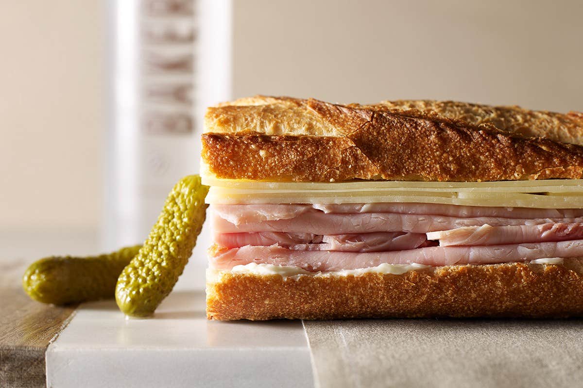 Close-up of a ham and cheese sandwich on crusty bread with pickles in the background.