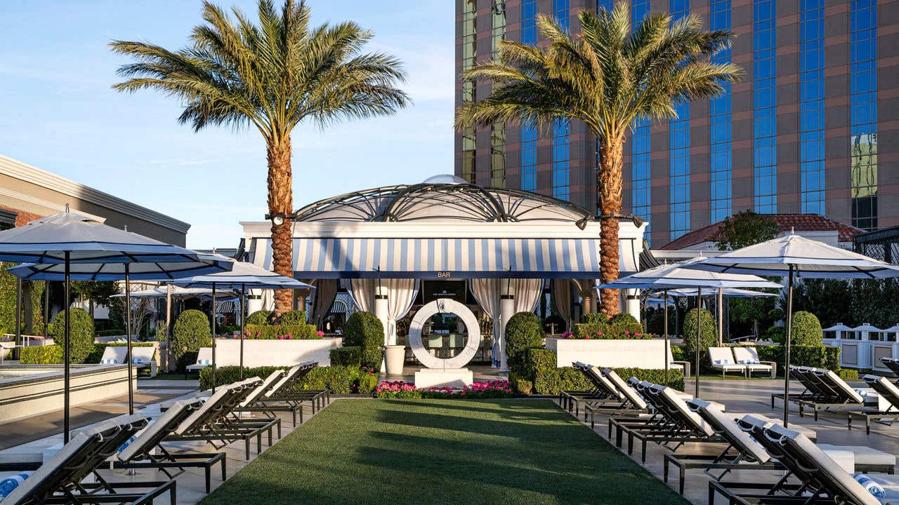 Outdoor lounge area with rows of chairs facing a central cabana pavilion, framed by two palm trees and surrounded by umbrellas and greenery.