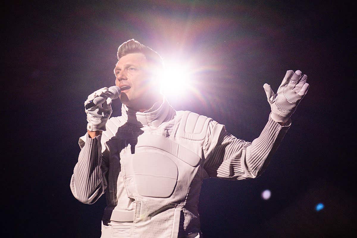 Nick Carter dressed in all white holding a microphone in front of a dark background with light shining from behind him. 