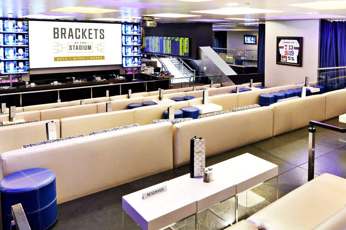 Modern sports lounge with large screen displays, stadium-style seating, and individual tables overlooking a central bar area.