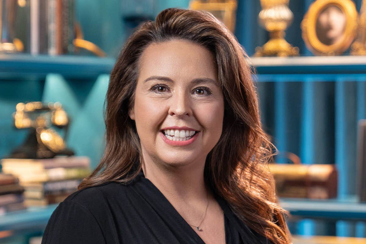 A woman with long, brown hair smiling in front of shelves with books and vintage decor pieces.