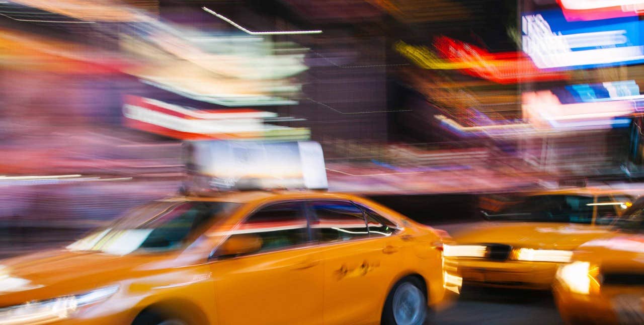 Blurred motion image of yellow taxis driving through a vibrant, neon-lit cityscape at night.