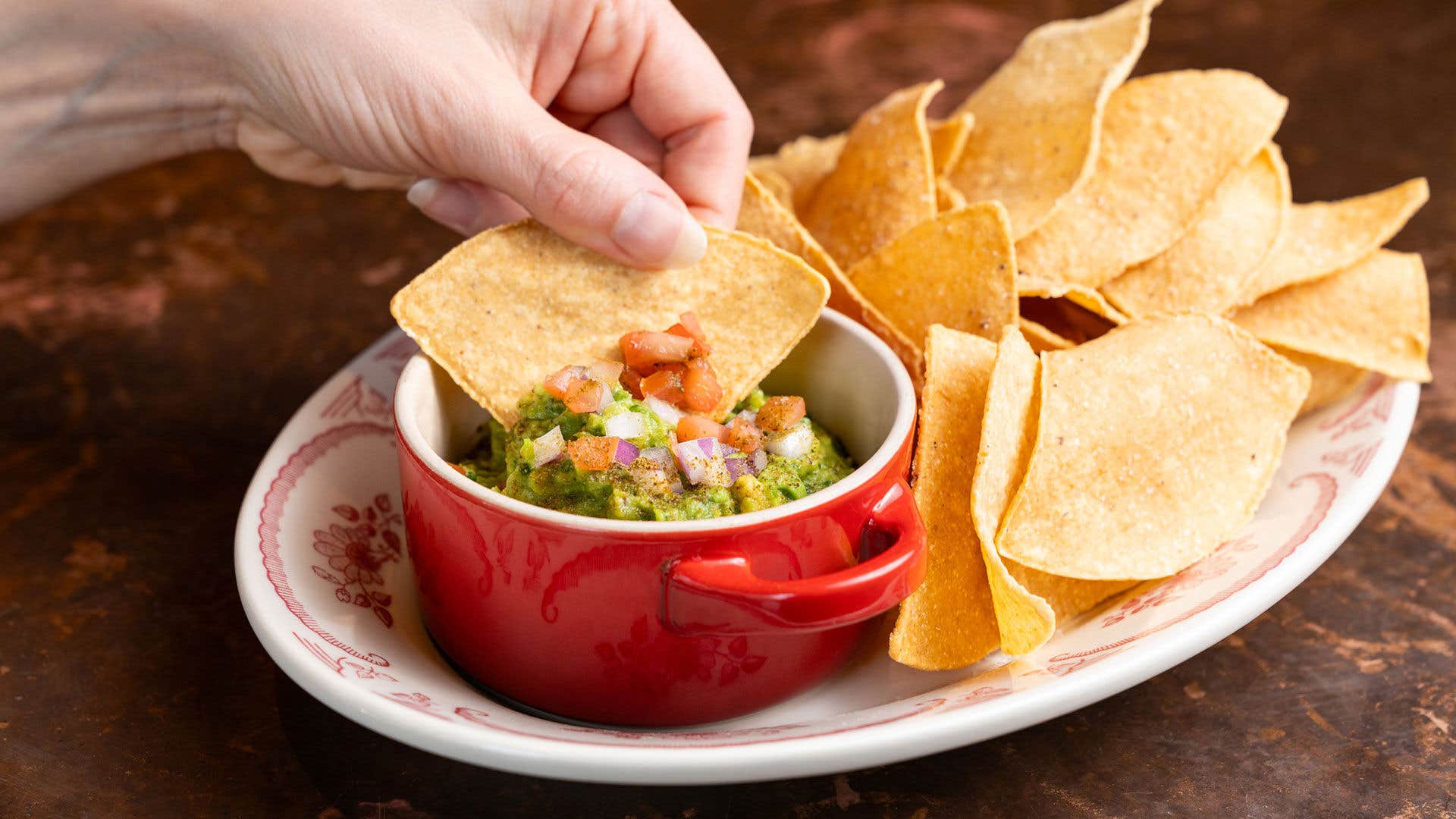 A hand holding a tortilla chip above a bowl of guacamole with additional chips on a plate.