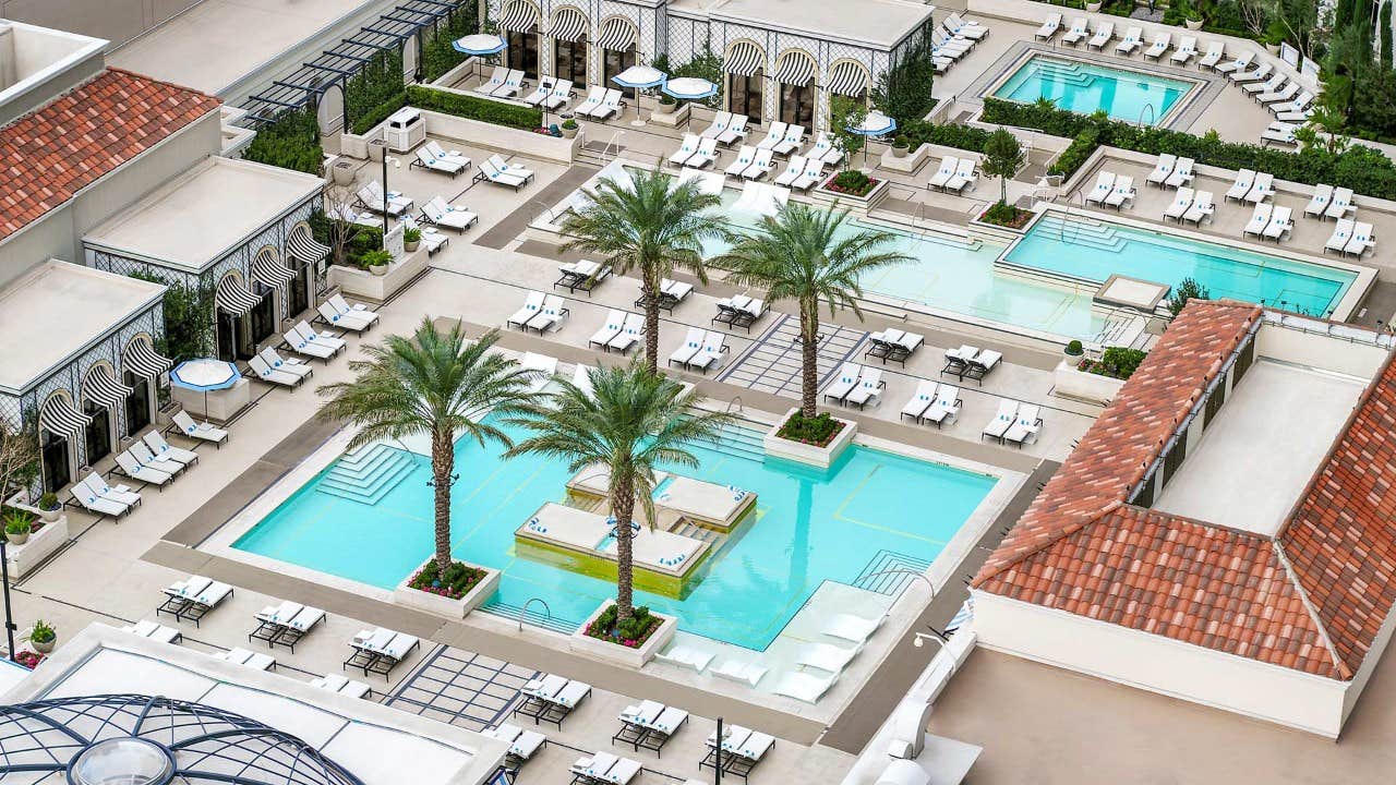 Aerial view of a luxurious pool area with multiple pools, lounge chairs, and palm trees surrounded by elegant buildings.