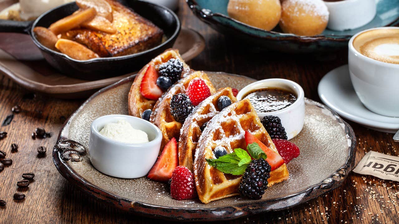 Waffles with berries, syrup, and cream on a rustic plate, accompanied by coffee and pastries in the background.