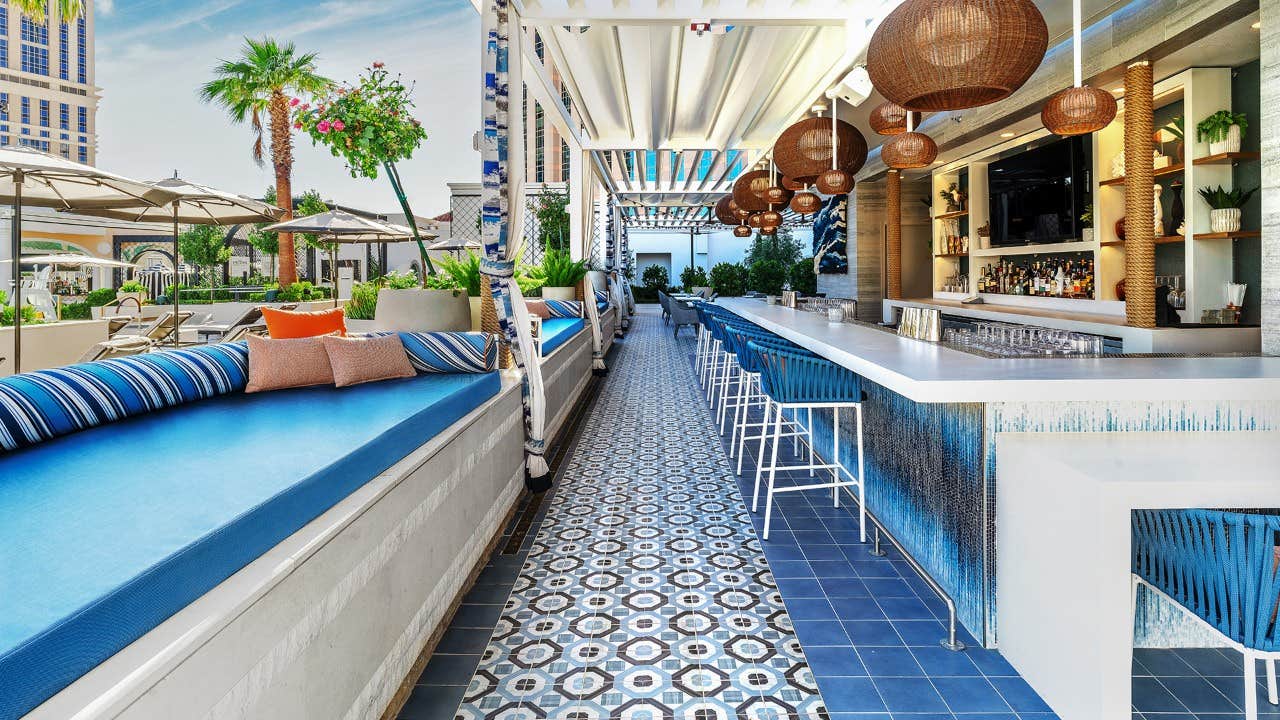 Outdoor bar and lounge area with patterned floor tiles, blue cushioned seating, hanging lamps, and tropical plants.