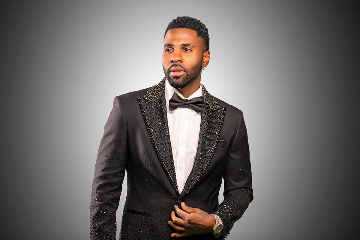 A man in a black suit with a bow tie stands against a plain grey background, looking off to the side.