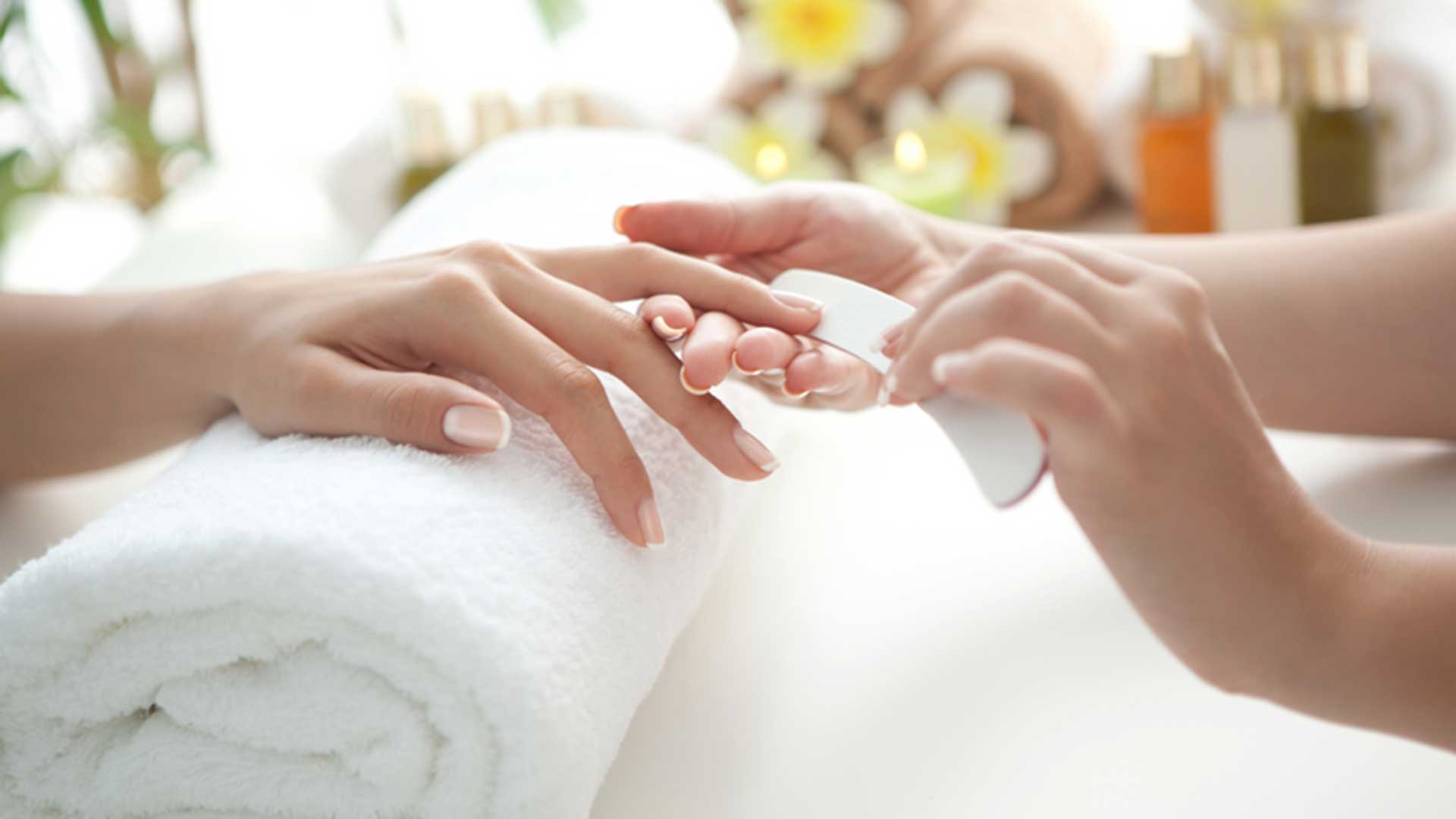 A person receives a manicure as another individual files the fingernails over a rolled white towel in a spa-like setting.