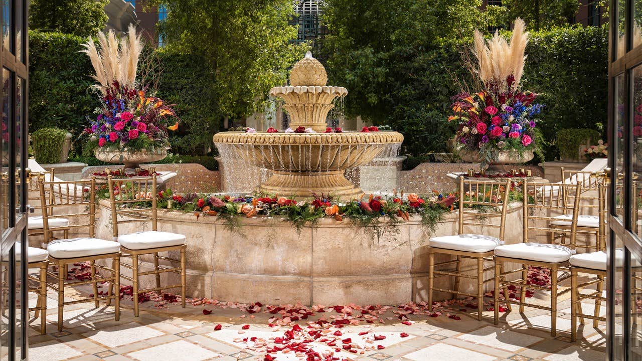 Elegant outdoor wedding setup with a stone fountain, floral arrangements, gold chairs with white cushions, and rose petals.