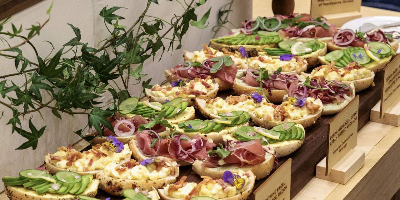 Open-faced bagel sandwiches topped with avocado, cured meat, egg salad, cucumbers, and edible flowers, arranged on wooden serving boards at a buffet.