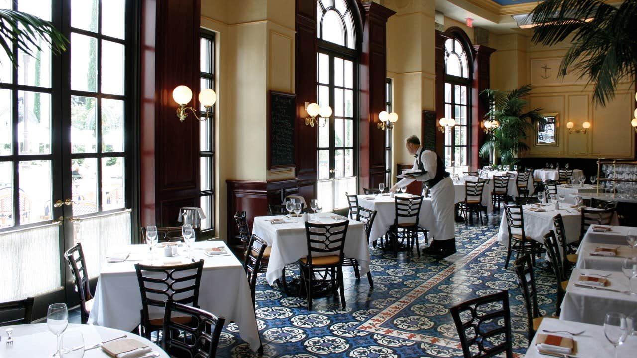 A server sets a table in a bright, elegant restaurant with large windows, white tablecloths, and patterned tile flooring.