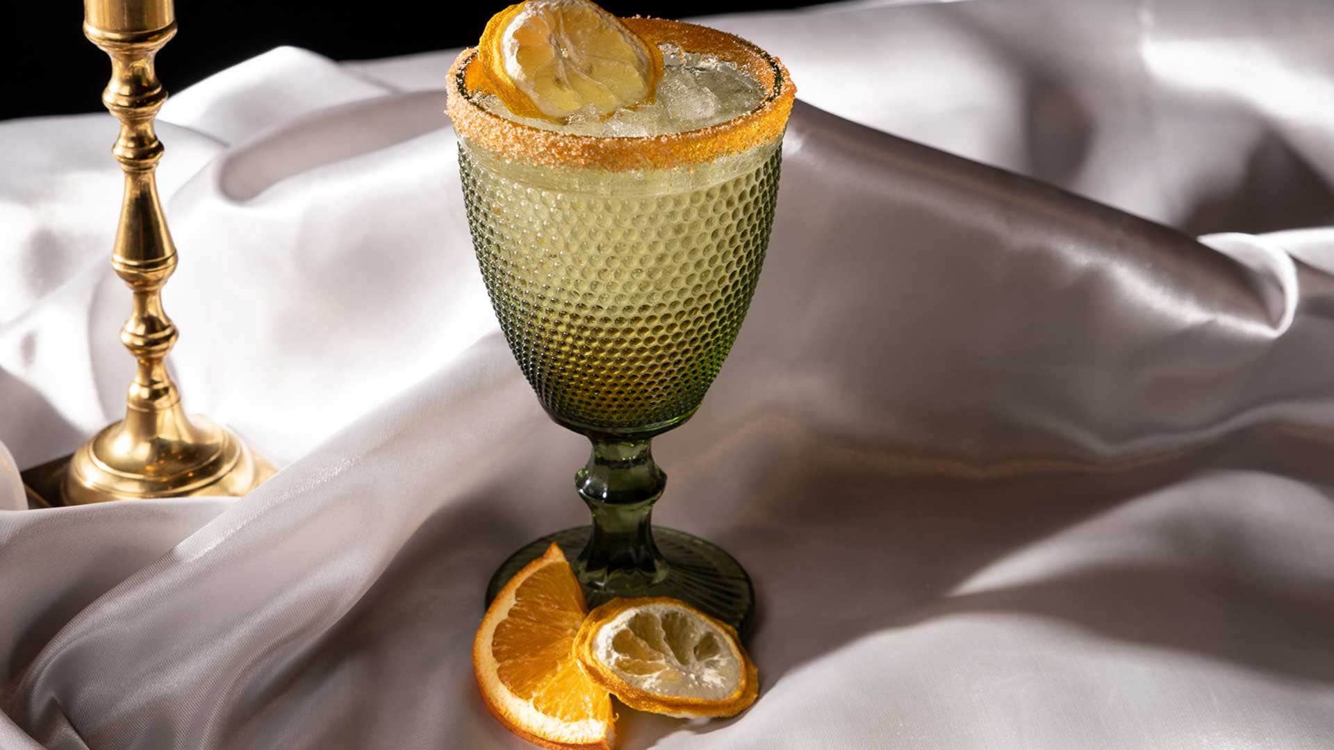 A green textured glass filled with a citrusy beverage, garnished with lemon and orange slices, on a silky fabric background.